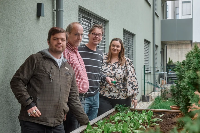Vier Personen stehen neben einem bepflanzten Hochbeet vor einem Wohnhaus, schauen in die Kamera und wirken stolz auf ihre gemeinsame Gartenarbeit.