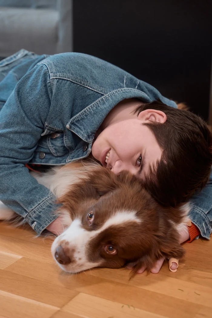 Eine Jugendliche mit kurzen dunkelbraunen Haaren liegt mit ihrem braun weißen Australian Shepherd auf einem Pakettboden und lächelt in die Kamera.