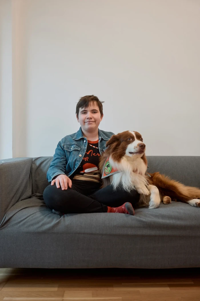 Eine Jugendliche mit kurzen dunkelbraunen Haaren sitzt mit ihrem braun weißen Australian Shepherd auf einem graue Sofa und schaut in die Kamera.