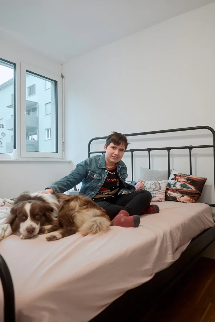 Eine Jugendliche mit kurzen dunkelbraunen Haaren sitzt mit ihrem braun weißen Australian Shepherd auf einem Bett und schaut in die Kamera.