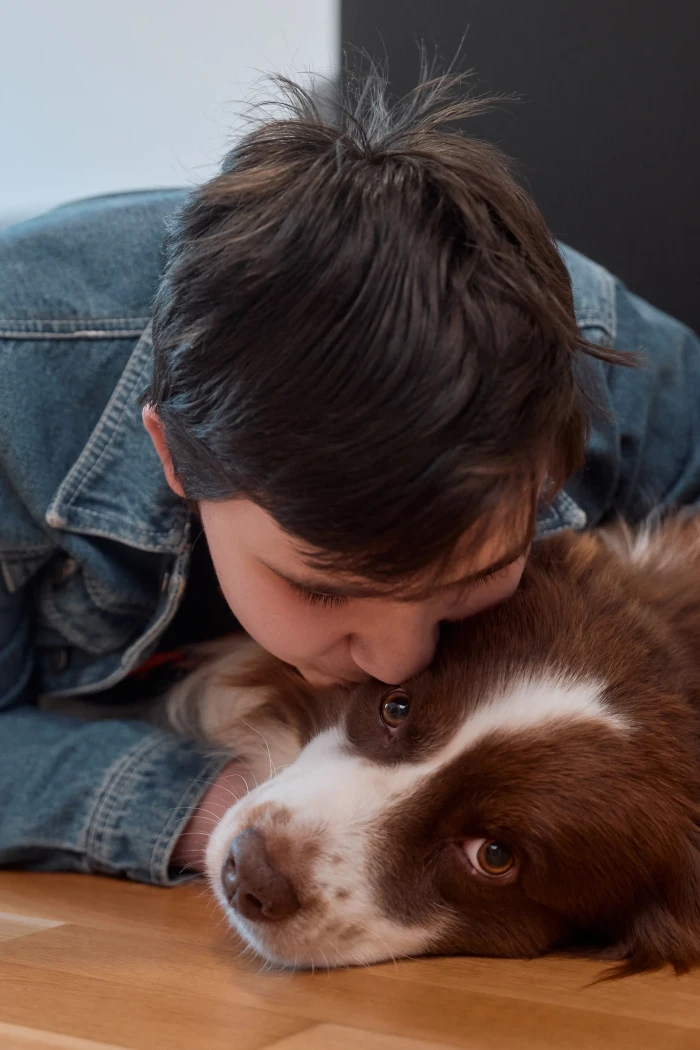 Eine Jugendliche mit kurzen dunkelbraunen Haaren liegt mit ihrem braun weißen Australian Shepherd auf einem Boden und gibt ihm einen Kuss auf die Wange.