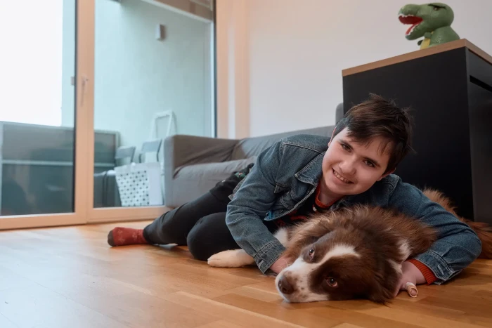 Eine Jugendliche mit kurzen dunkelbraunen Haaren liegt mit ihrem braun weißen Australian Shepherd auf einem Boden und lächelt in die Kamera.