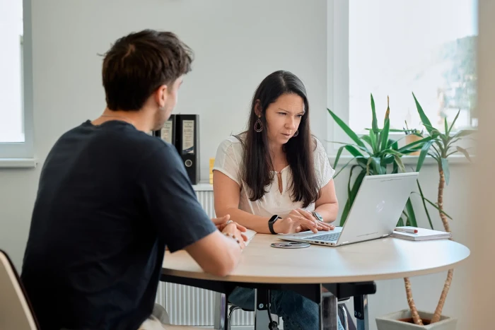 Eine Frau und ein Mann sitzen sich gegenüber an einem Tisch, die Frau sieht in ihren Laptop rein.